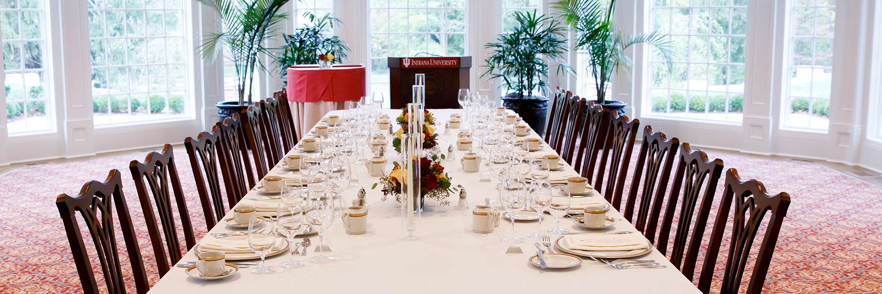 A long table is set with china and stemware for a formal dinner; a podium with IU sign stands at the head of the table.