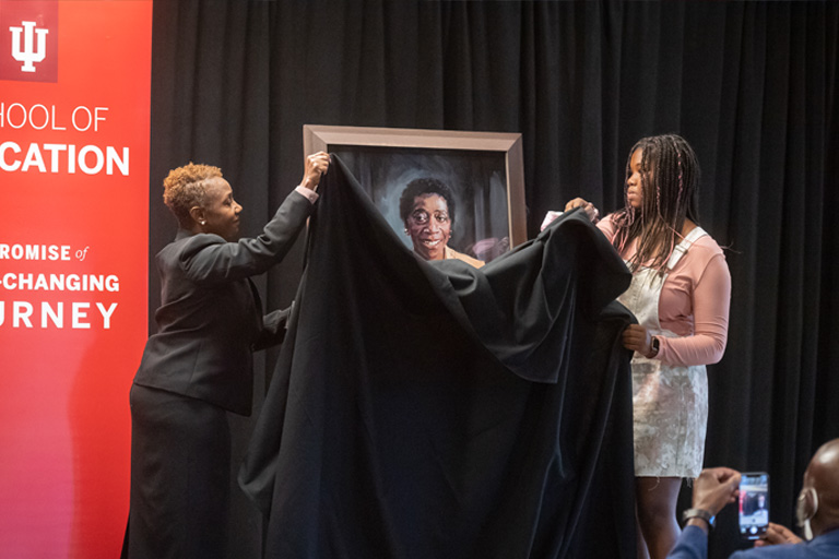 Two women unveil an oil painting covered by black velvet drape in a formal IU ceremony. 