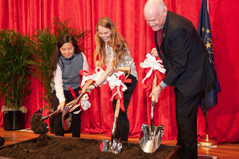 Two women and a man turn soil over with ceremonial shovels in a groundbreaking ceremony. 