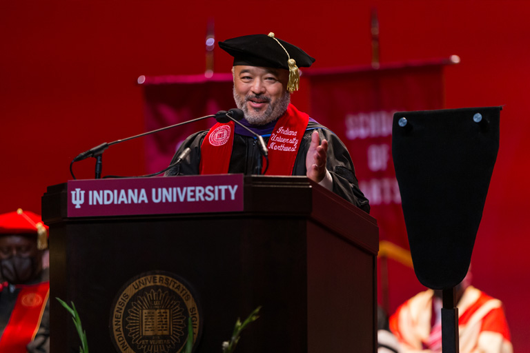 A man in formal academic attire speaks warmly, gesturing from the podium.