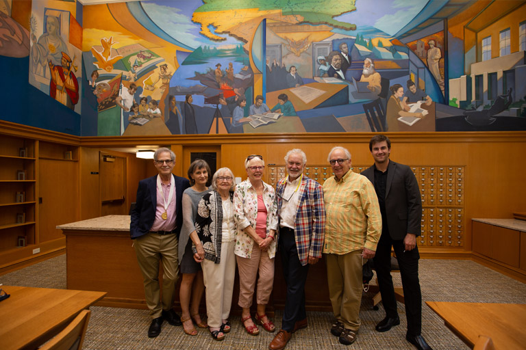 A group of people, the furthest left wearing a university medal, pose in front of a breathtaking large-format mural. 